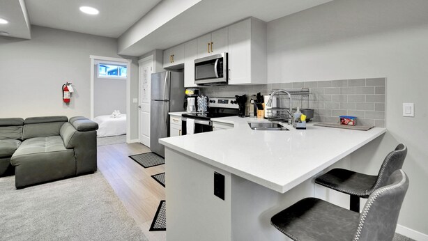 A modern kitchen with white cabinetry, a central island, and a grey sofa in a living area. - Family Retreat 2BDR 2BT / Remote Work Ready / Lake Access / Games / South Health (Calgary)