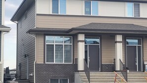 A two-story residential building with a brick foundation, large windows, and a front porch with steps. - Family Retreat 2BDR 2BT / Remote Work Ready / Lake Access / Games / South Health (Calgary)