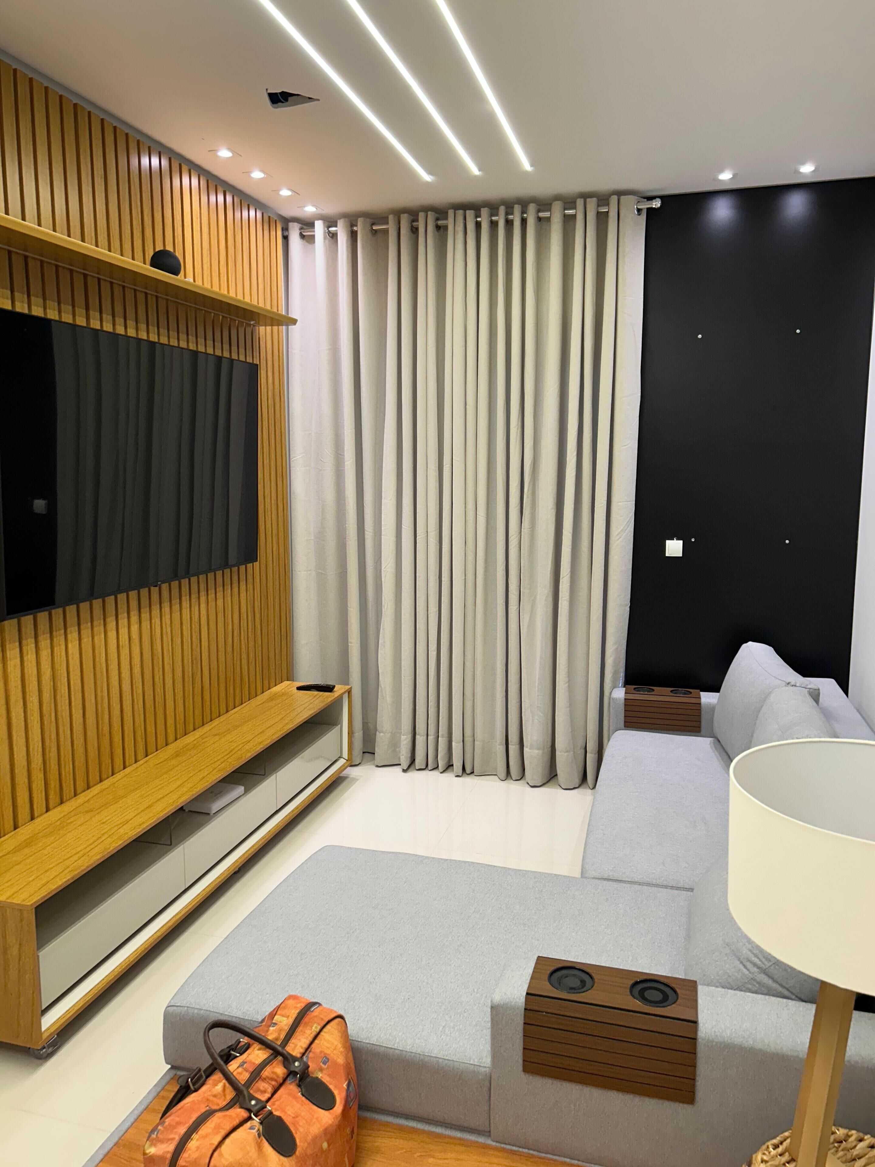 A modern living room with a flat-screen TV mounted on a wooden panel, a grey sofa, a small table with a lamp, and a grey curtain.