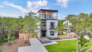 A modern two-story house with a balcony, surrounded by trees and a well-maintained lawn. - Lake Travis Retreat w/ Pool, Views & Outdoor Kitchen. Perfect for groups (Lago Vista)