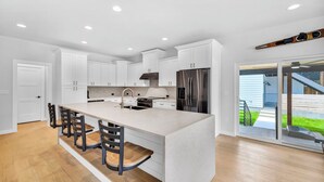 A modern kitchen with white cabinets, a large island with bar stools, stainless steel appliances, and a view to a backyard. - Lake Travis Retreat w/ Pool, Views & Outdoor Kitchen. Perfect for groups (Lago Vista)