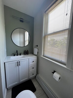 Une salle de bain avec un meuble-lavabo blanc, un miroir rond, une fenêtre avec des stores et un porte-rouleau de papier toilette.
