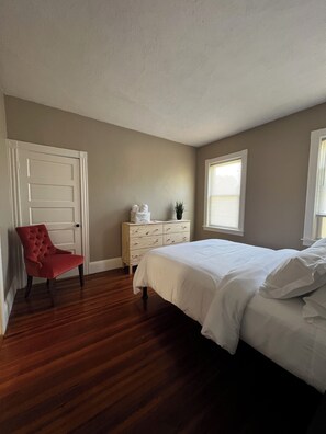 Une chambre avec un plancher en bois, un lit avec une literie blanche, une chaise rouge, une commode et une fenêtre avec des stores.