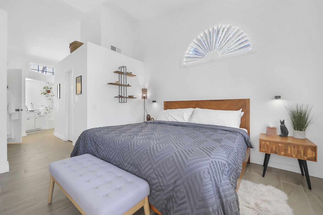 Quarto moderno com uma cama grande, mesa de cabeceira de madeira, um banco e um ventilador na parede.