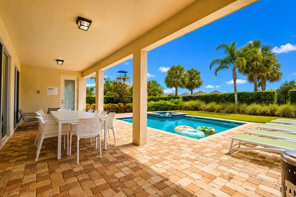 A covered outdoor area with a pool, lounge chairs, and a table with chairs.