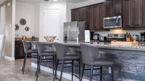 A modern kitchen with dark wood cabinets, a stainless steel refrigerator, a microwave, and a granite countertop. There are three bar stools and a decorative mirror on the wall. - Stunning 9BR/8.5BA Retreat with Private Pool & SPA-280CW (Kissimmee)