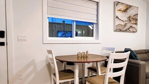 Un comedor con una mesa redonda y cuatro sillas, una ventana con persianas, una obra de arte colgada en la pared y un sofá de color oscuro.