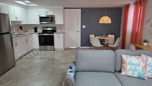 Eine moderne Küche mit weißen Schränken, einem Edelstahl-Kühlschrank, einer Mikrowelle und einem Ofen. Es gibt einen Essbereich mit Tisch und Stühlen, sowie ein graues Sofa mit Kissen im Strandstil.