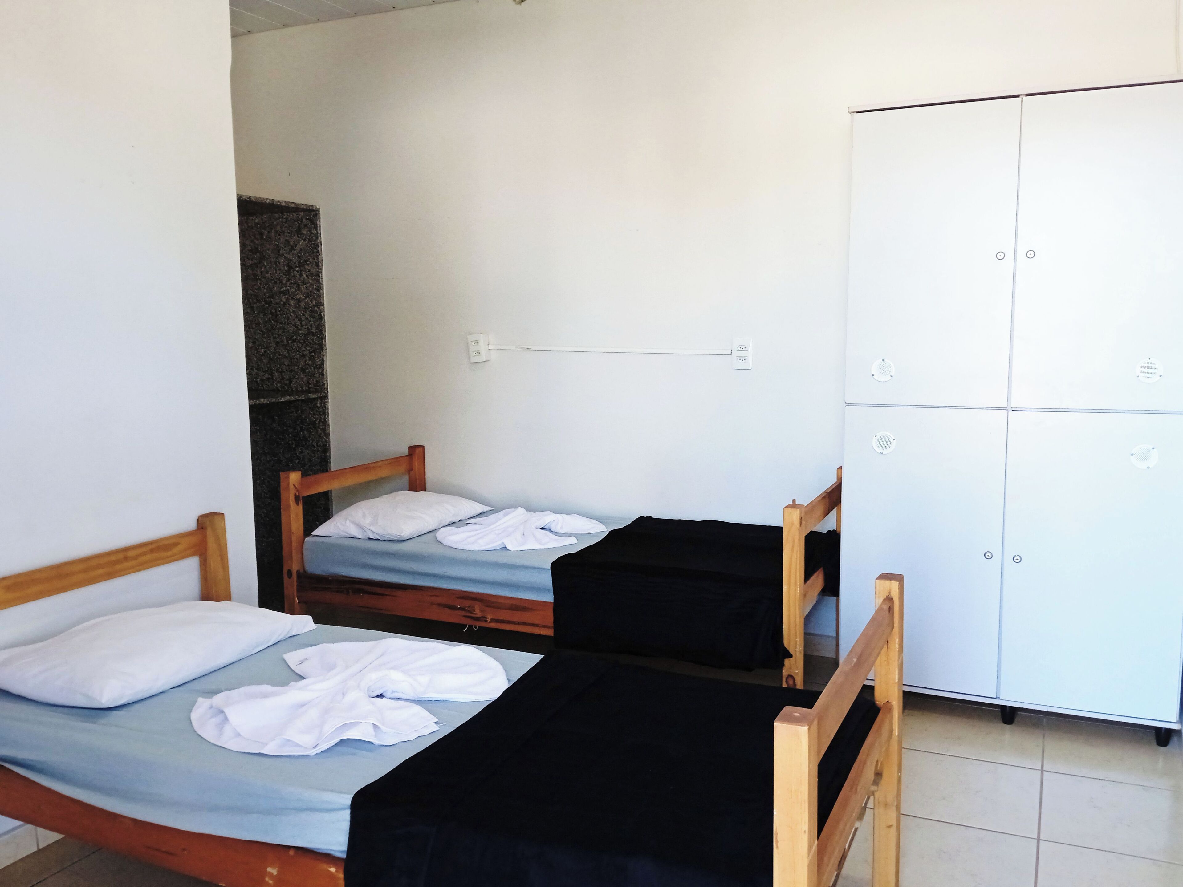 Une pièce avec deux lits en bois, l’un avec un matelas bleu et l’autre avec un matelas noir. On y trouve une armoire blanche à quatre portes et une petite étagère.