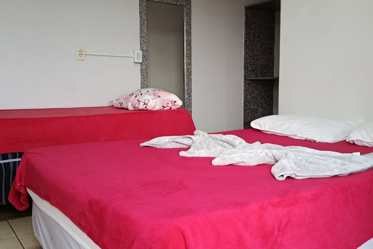 Une chambre avec deux lits, l’un recouvert d’une couverture rouge et l’autre d’une couverture blanche. Il y a un miroir au mur et une petite étagère.
