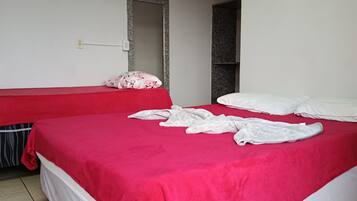 Quarto com duas camas, uma com colcha vermelha e outra com colcha branca. Há um espelho na parede e uma pequena prateleira.