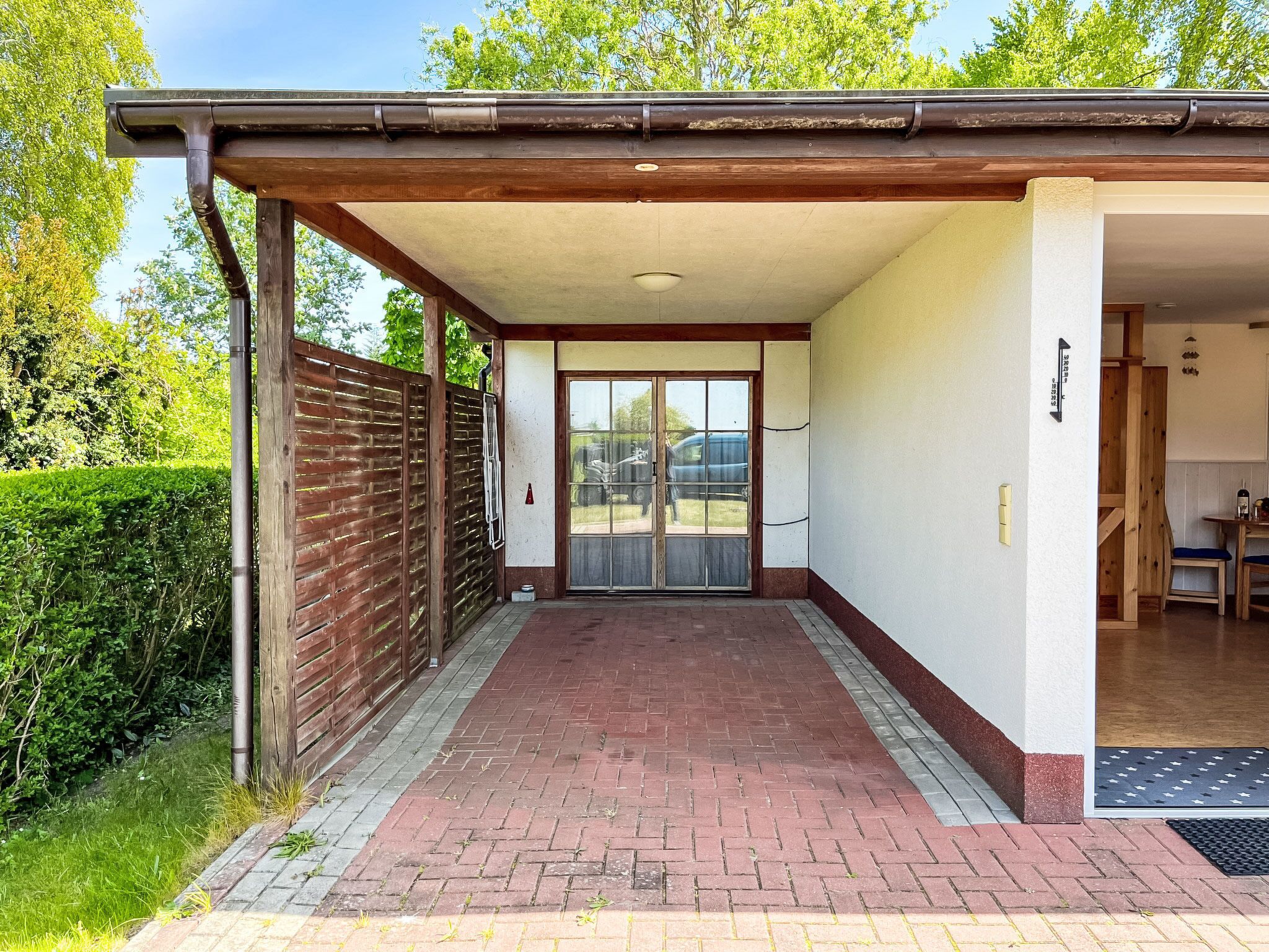 A covered outdoor area with a brick floor, a wooden fence, and a door leading to a room.