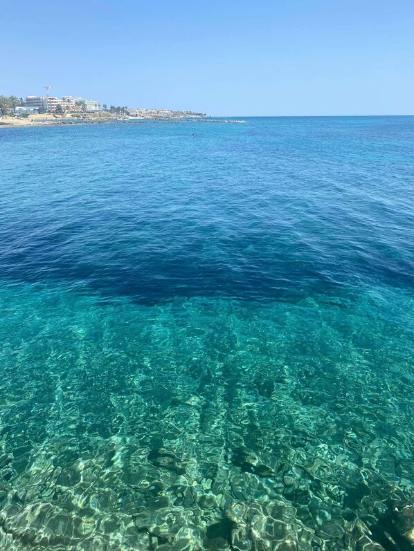 A coastal view with clear blue water and a distant building complex.