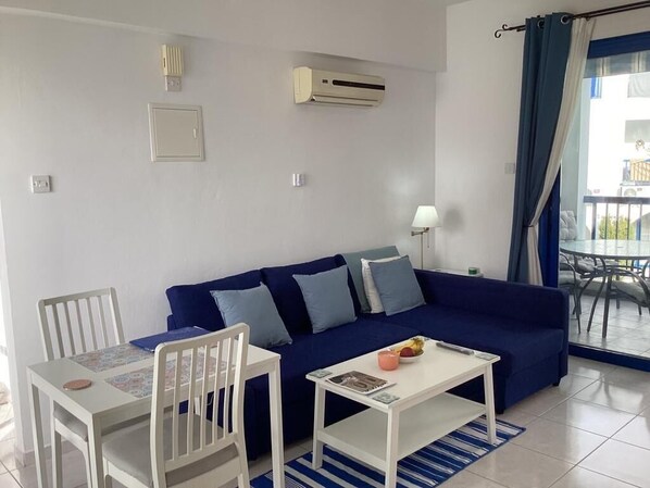 A living room with a blue sofa, white chairs, a small table, and an air conditioning unit on the wall.