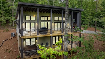 Une cabane à deux étages dotée d’une terrasse, de grandes fenêtres et d’une cheminée.