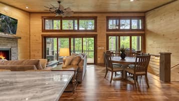 A spacious living area with a fireplace, a dining table with chairs, and a large window overlooking a forested area.