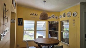 Un coin repas avec une table ronde en bois et quatre chaises, un luminaire suspendu et un mur décoré d’assiettes et d’une enseigne sur laquelle on peut lire : « CUISINE, lieu de rencontre ».