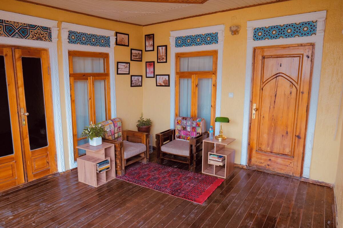 A cozy room with wooden floors, two wooden doors, a small table with a plant, two chairs, a rug, and a wall decorated with framed pictures.