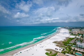 An aerial view of a beach resort with a long stretch of white sand, turquoise waters, and a mix of beach umbrellas and buildings. - Charming 2-bedroom condo walking distance to Siesta Key’s beach! (Siesta Key)