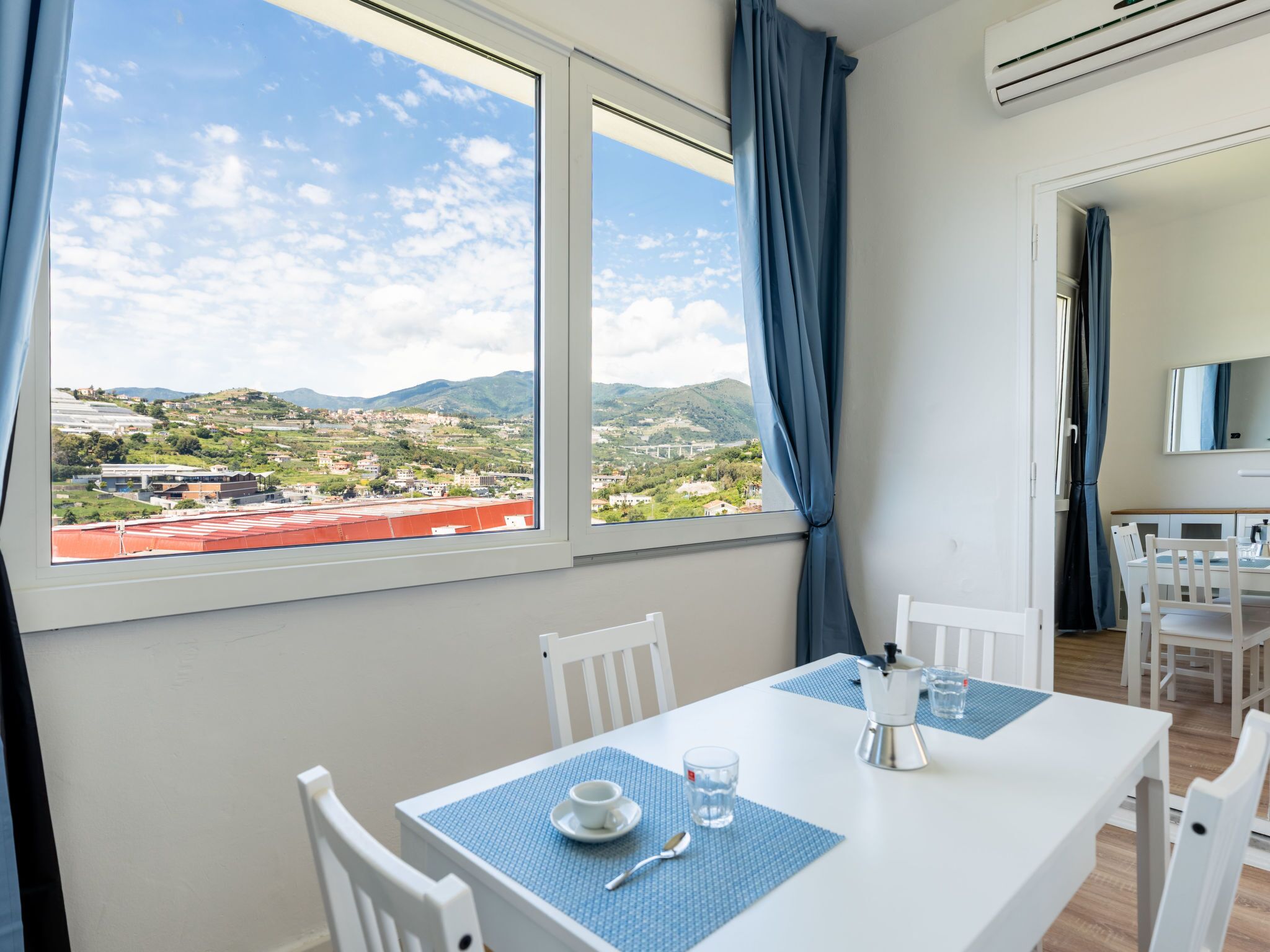 Ein Zimmer mit Esstisch, Stühlen, einer Kaffeemaschine und Blick auf eine Berglandschaft durch große Fenster.