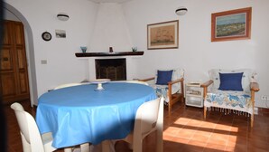 A dining area with a round table covered by a blue tablecloth, surrounded by white chairs. There is a fireplace with a mantel above the table, and a framed painting on the wall to the right. - Villa with large garden (Marina di Campo)