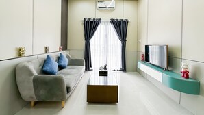 A modern living room with a grey sofa, blue cushions, a glass coffee table, a flat-screen TV on a teal stand, and a window with black curtains. - Golden Sun Hotel Apartments 5 (Ho Chi Minh City)