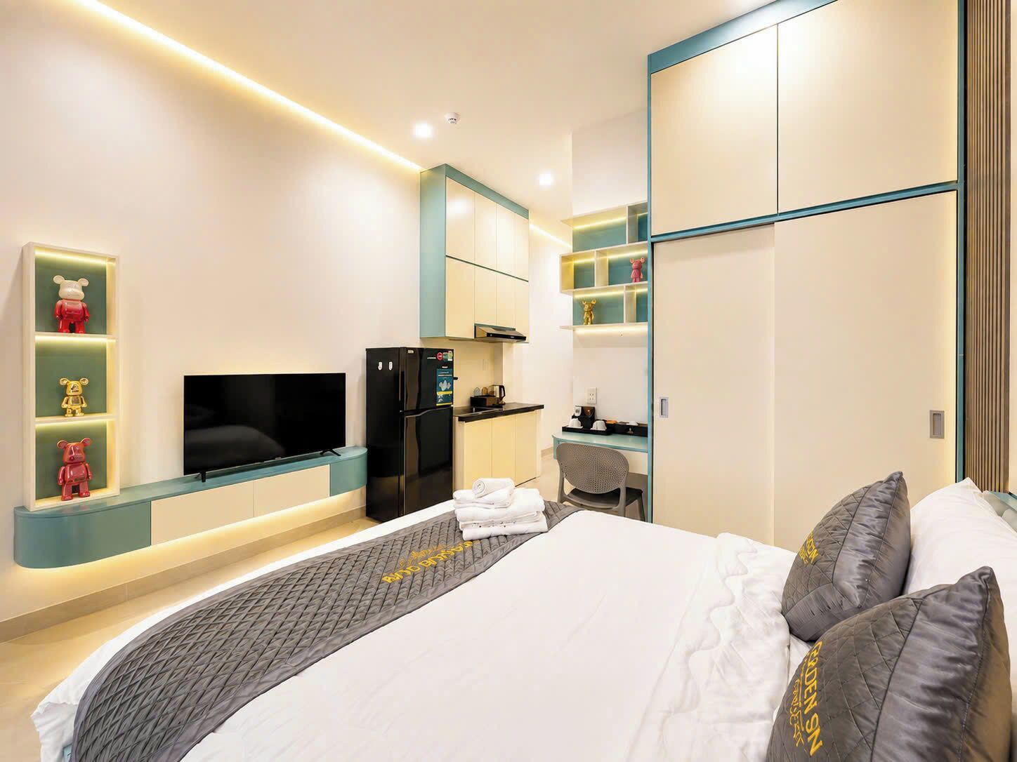 A modern hotel room with a bed, a TV, a refrigerator, a desk, and a kitchenette.