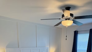 A neatly made bed with white bedding and gray pillows, a white nightstand with a lamp, and a ceiling fan.