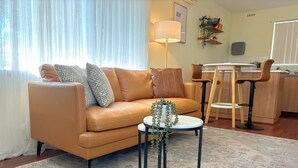 Ein Wohnzimmer mit einer Couch, einem kleinen runden Tisch mit einer Pflanze, einem Essbereich mit Tisch und Stühlen sowie einer Küchenzeile mit Spüle und Arbeitsfläche.