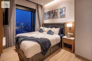 Habitación de hotel con una cama grande, una mesita de noche con lámpara y vistas a la ciudad al anochecer.