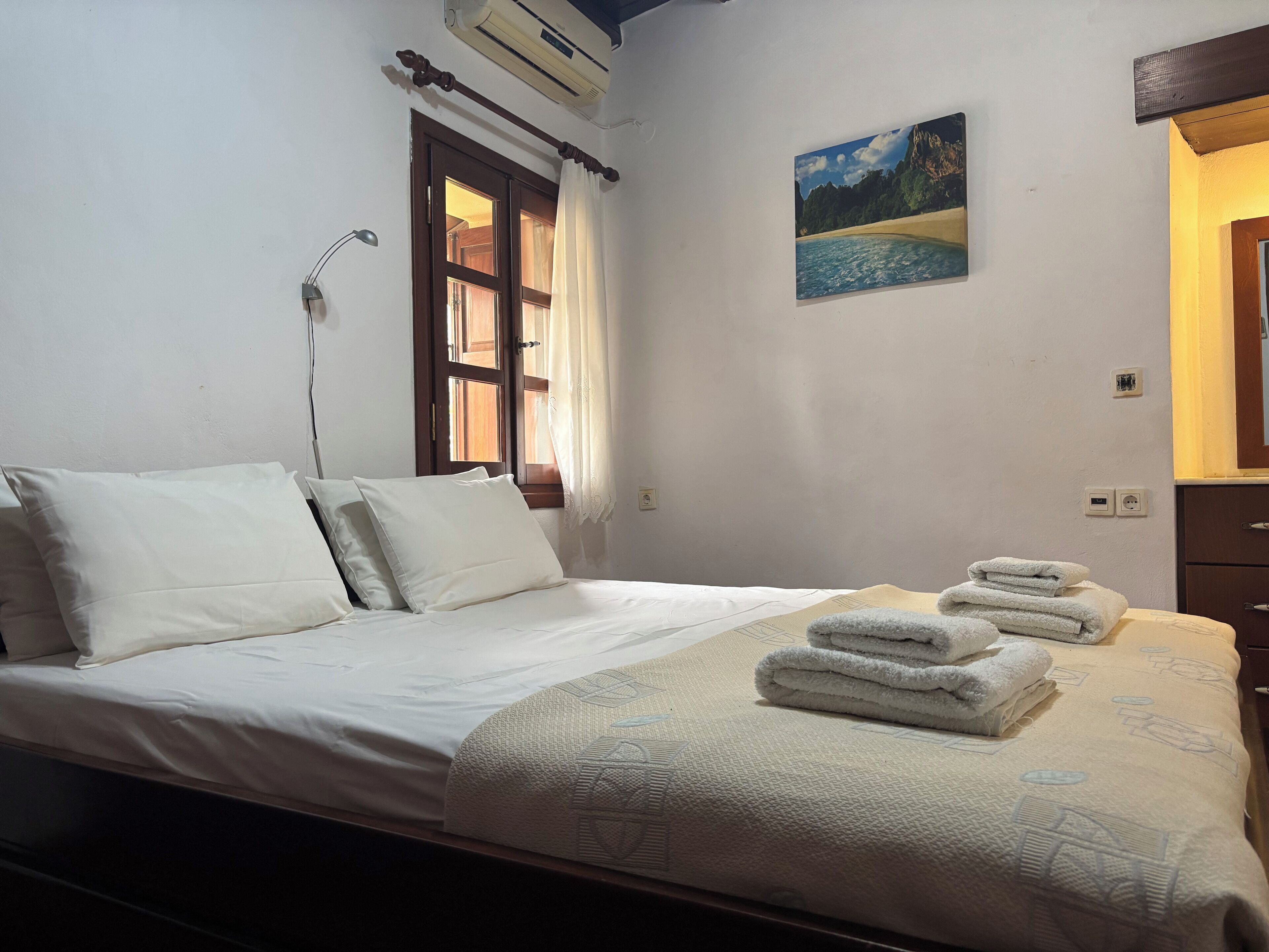 A neatly made bed with pillows and a blanket, a window with curtains, a painting on the wall, and a stack of towels on the side.