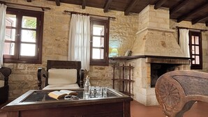 A rustic room with a stone wall, wooden ceiling beams, a fireplace, a glass-top table with books and glasses, and a wooden chair. - Melina Stone Villa (Roumelí)