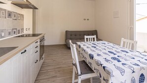 A compact kitchen with a built-in stove, white cabinets, and a grey sofa in a dining area with a table covered in a fish-patterned tablecloth. - Il Gelso - Sea View Holiday Home in Pomonte - Elba Island (Pomonte)