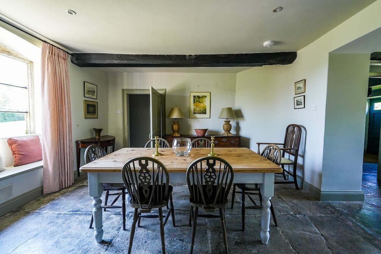 A dining room with a wooden table and chairs, a window with curtains, and a stone floor.