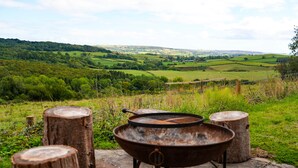A rustic outdoor cooking setup with a metal pot on a stand, surrounded by wooden stumps, overlooking a green valley with rolling hills. - Spacious Country Farmhouse in Rewilding Landscape (Mapperton)