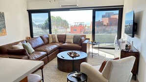 A modern living room with a brown leather sectional sofa, a round coffee table, a white armchair, and a TV mounted on the wall. - Surf Paraiso @The Point (Los Cerritos, El Pescadero)