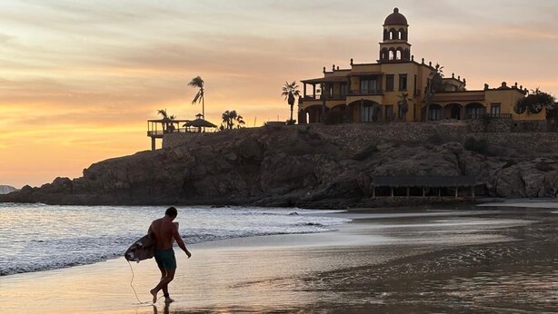 A beach scene with a person carrying a surfboard, a building with a dome and arched windows, and a rocky shoreline. - Surf Paraiso @The Point (Los Cerritos, El Pescadero)