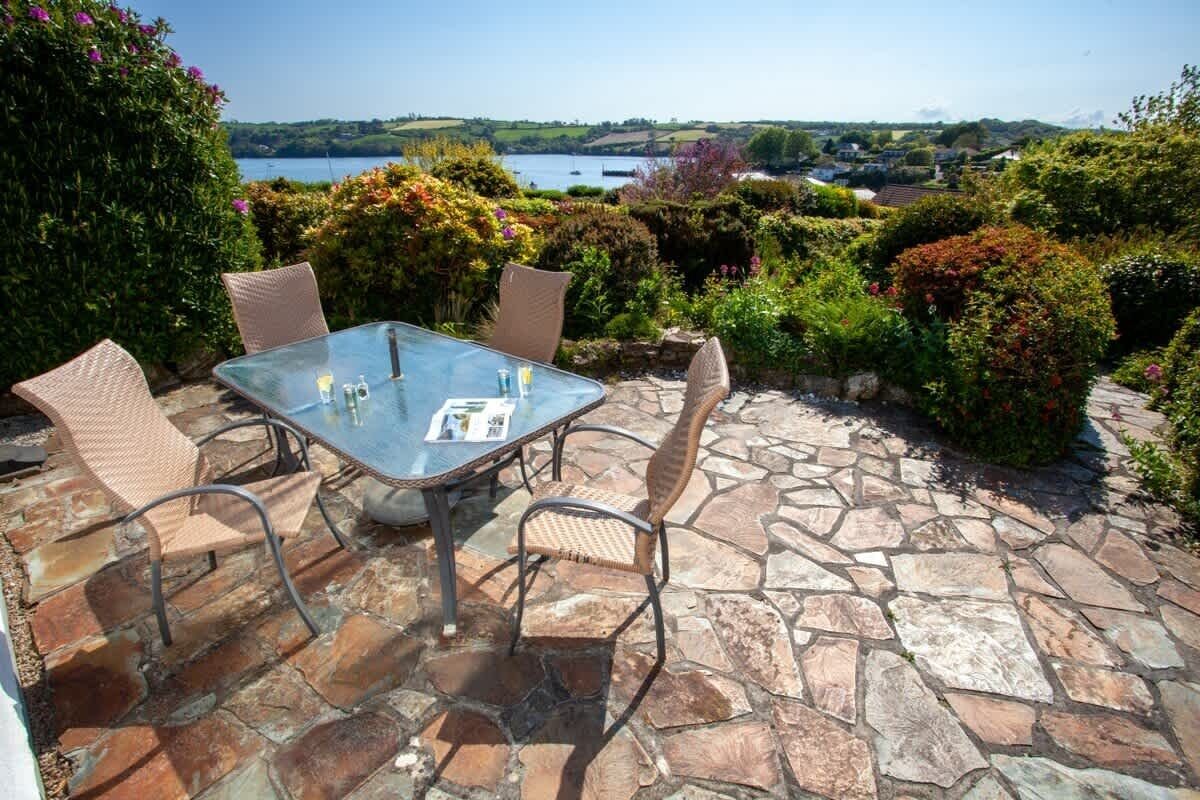 An outdoor patio with a glass table and wicker chairs, surrounded by stone flooring and lush greenery.
