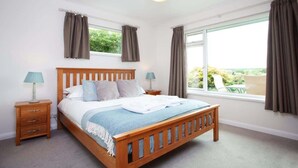 Un dormitorio con cama de madera, dos mesitas de noche con lámparas, un ventanal con cortinas y vistas a zonas verdes.