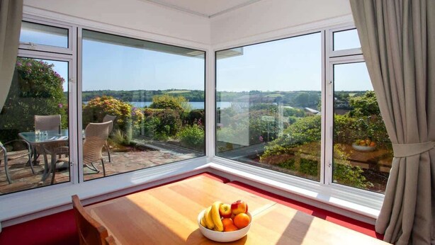 Una habitación con un ventanal grande que da a un jardín y un lago. Hay una mesa de madera con un frutero y una silla.