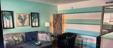 A living room with a sofa, a coffee table, a chair, and a small table with a lamp. There are framed pictures on the wall and a window with blinds.