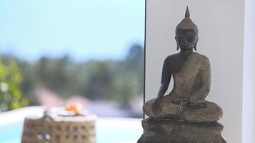 Une statue représentant une figure assise sur un piédestal, placée sur une table en verre près d’une fenêtre donnant sur un paysage extérieur flou.