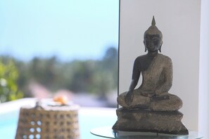 Eine Statue einer sitzenden Figur auf einem Sockel, platziert auf einem Glastisch in der Nähe eines Fensters mit unscharfem Blick ins Freie.
