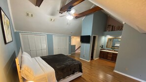 A bedroom with a bed, a nightstand with a lamp, a ceiling fan, and a view of a bathroom and kitchen.