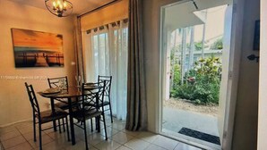 A dining area with a table and chairs, a painting on the wall, and a view of a garden through a sliding door.