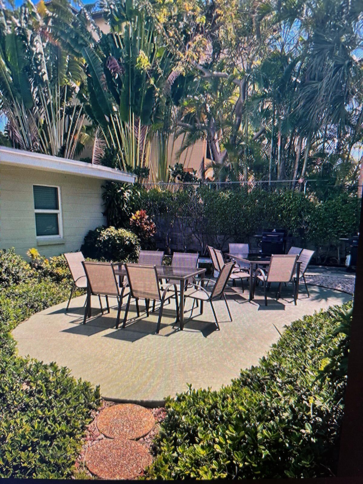 An outdoor patio with a table and chairs, surrounded by lush greenery and a house in the background.