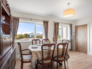 A dining area with a table set for four, wooden chairs, a chandelier, and large windows with curtains. - Atlantic Haven Water View (Cashel)
