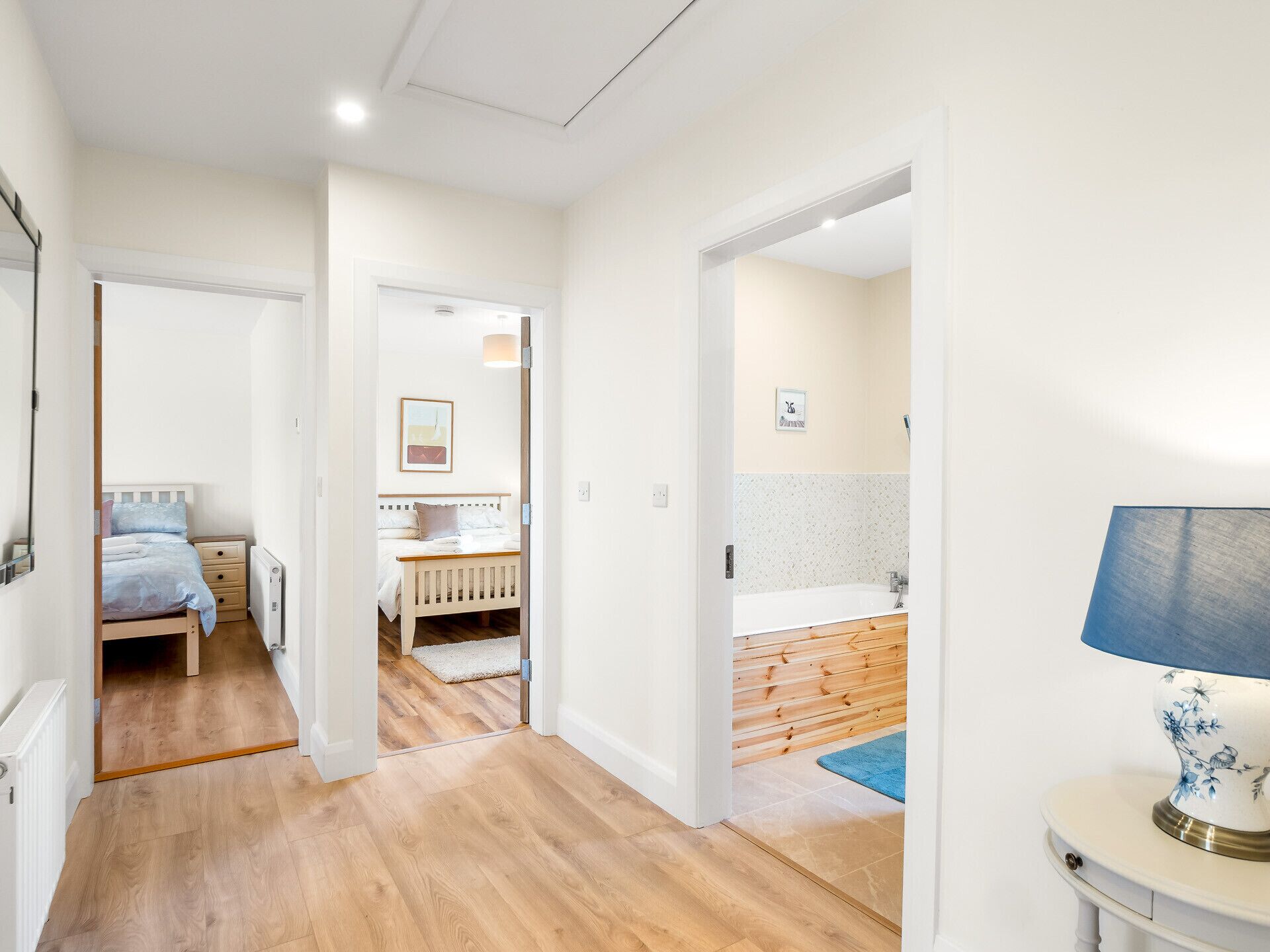 A bedroom with a bed, bedside table, and a nightstand. There is a bathroom with a bathtub, sink, and mirror. The room has wooden flooring and white walls.