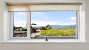Janela com vista para área gramada, caminho e corpo d'água com montanhas ao longe.