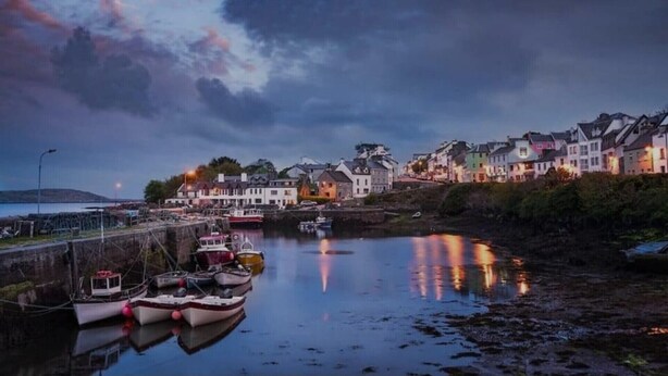 Uma vila costeira ao entardecer, com barcos atracados na margem e edifícios cujas luzes se refletem na água.